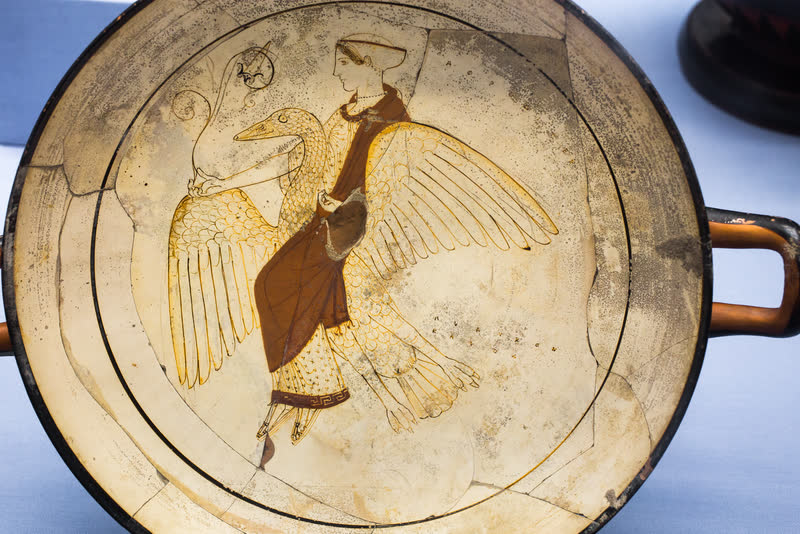 Afrodite sentada de través sobre um grande ganso de asas abertas, o manto escuro sobre o vestido e o nome dela escrito no campo, ao lado da cabeça. Interior de uma taça (kylix) ática de fundo branco, ca. 460 a.C., atribuída ao Pintor de Pistoxenos. Museu Britânico, Londres (1864,1007.77).