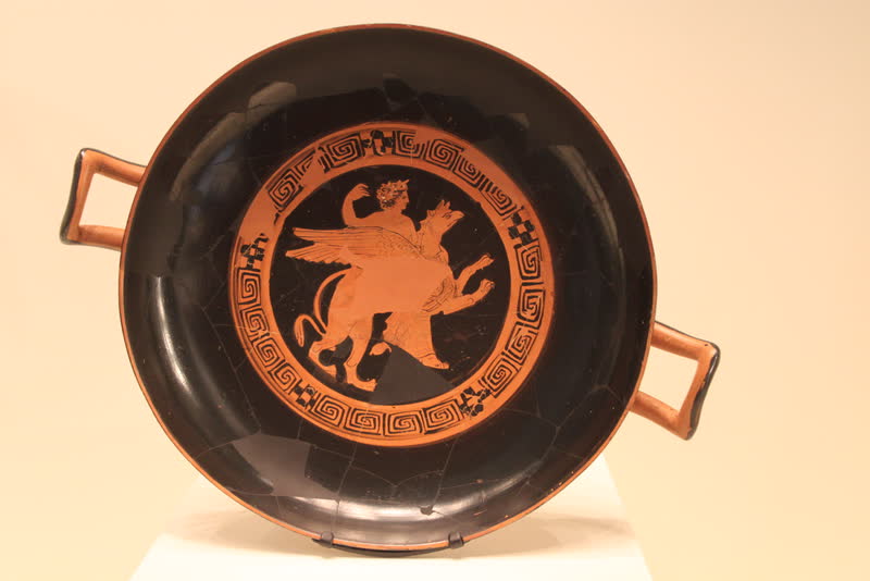 Apolo sentado de lado sobre um grifo de asas abertas, o braço erguido e o manto caindo sobre o dorso do bicho; em volta, a coroa de meandros e xadrez. Interior de uma taça (kylix) ática de figuras vermelhas, ca. 380 a.C. Getty Villa, Los Angeles.