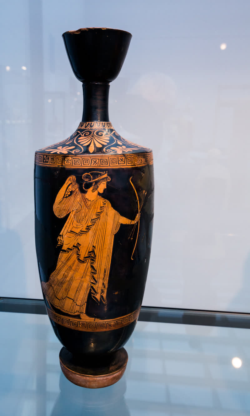 Ártemis caminha para a direita segurando o arco e duas flechas na mão esquerda e tirando outra da aljava com a direita. Lécito ático de figuras vermelhas, ca. 480–470 a.C., de pintor não atribuído; 43,6 cm de altura. Staatliche Antikensammlungen, Munique (NI 7514).