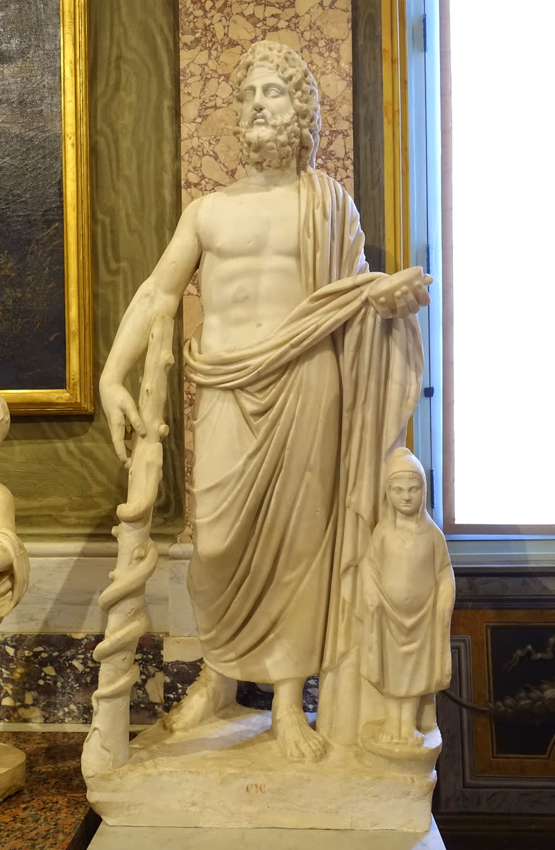 Asclépio de manto sobre o ombro, apoiado no bastão em que se enrola a serpente; ao lado, encapuzado, o pequeno Telésforo, o daimon da convalescença. Estátua romana em mármore, século II d.C. Galleria Borghese, Roma.