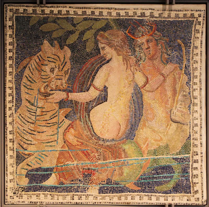 Uma nereida cavalga o monstro marinho de corpo listrado e cauda enrolada, segurando-o pelo focinho; atrás dela, um tritão de cauda verde. Mosaico romano. Musée des Beaux-Arts, Lyon.