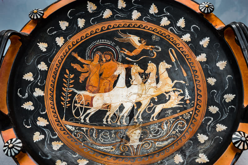 Hades arrebata Perséfone na quadriga, com Eros voando à frente e um cão correndo entre os cavalos; embaixo, uma cabeça feminina sai de um ornamento vegetal. Interior de um podanipter apuliano de figuras vermelhas, ca. 330–320 a.C., do Pintor do Mundo Inferior. Staatliche Antikensammlungen, Munique.