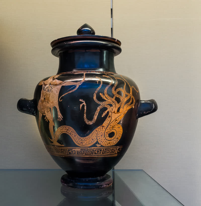 A Hidra de Lerna enrolada sobre si, o corpo escamado e um leque de pescoços terminando cada um em sua cabeça; à esquerda, Héracles avança sobre ela. Face A de um estâmnos ático de figuras vermelhas com tampa, ca. 480–470 a.C., atribuído ao Pintor de Géras. Museu Britânico, Londres (1929,0513.1).
