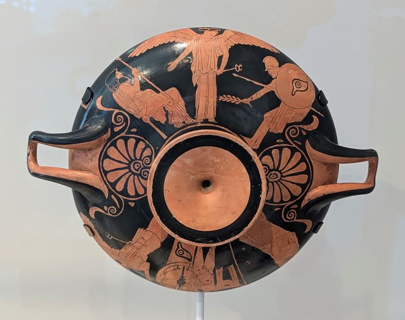 Íris, de asas abertas e cadúceu na mão, apresenta a Zeus sentado no trono um guerreiro de escudo e lança. Exterior de um cálice ático de figuras vermelhas, ca. 470 a.C., atribuído ao Pintor de Pentesileia. Harvard Art Museums · Arthur M. Sackler Museum, Cambridge.