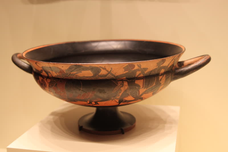 Caçadores a pé e a cavalo cercam o javali, de lanças em riste, num friso que dá a volta na taça. Cálice ático de figuras negras, ca. 560 a.C. Getty Villa, Malibu.