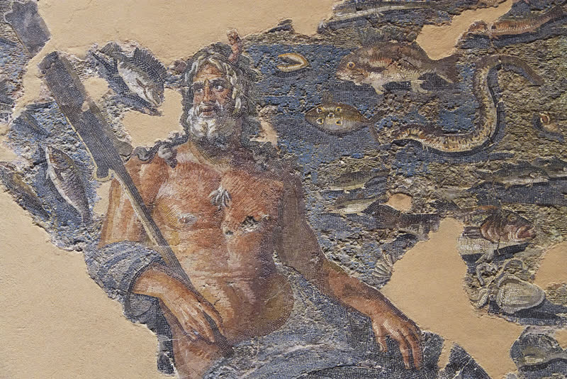 Oceano emerge de perfil entre peixes, enguias e conchas, as pinças de caranguejo sobre a testa e o remo apoiado no ombro. Mosaico romano de Oceano e Tétis (inv. 850), Museu de Arqueologia de Hatay, Antáquia.